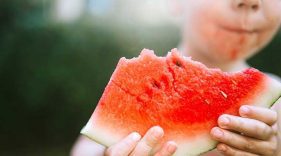 Kid holding out a large piece of watermelon that he is eating